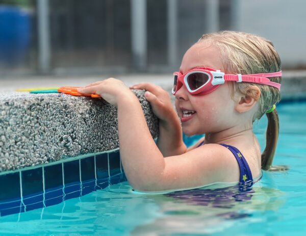 Little girl in goggles hangs on side of the pool