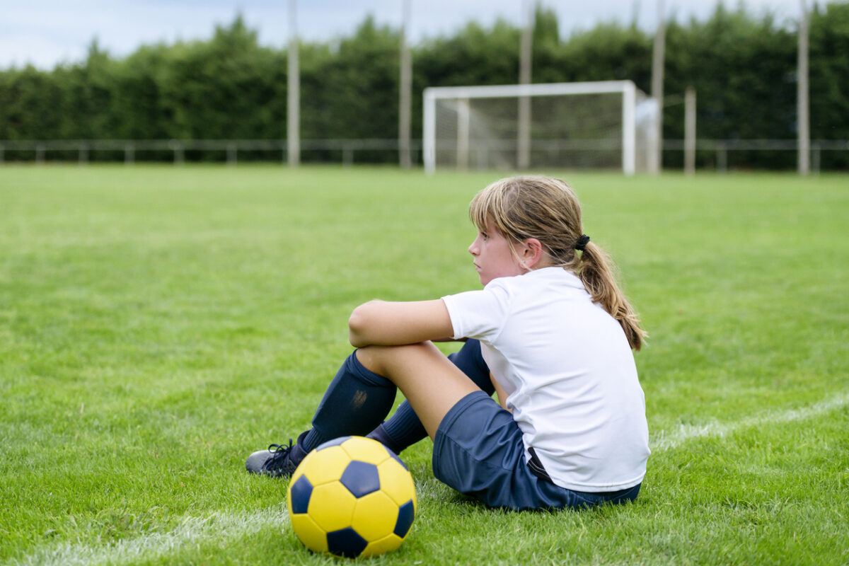 Girl in a soccer uniform sitting on the sidelines with a ball, reflecting after the game.