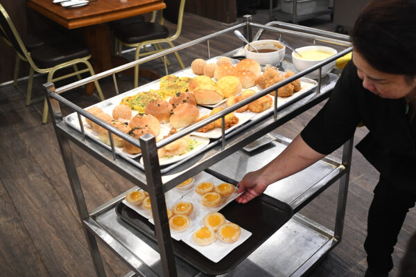 Woman leans over a rolling metal cart of dim sum, filled with multiple little trays of various items.