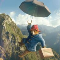 Paddington Bear flies through air with suitcase and umbrella, mountainscape in background