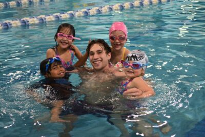 Adult instructor poses with four kids hanging off him in pool