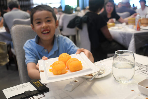 Smiling child holds out dish with bright orange treats to the camera, at the table of a restaurant.