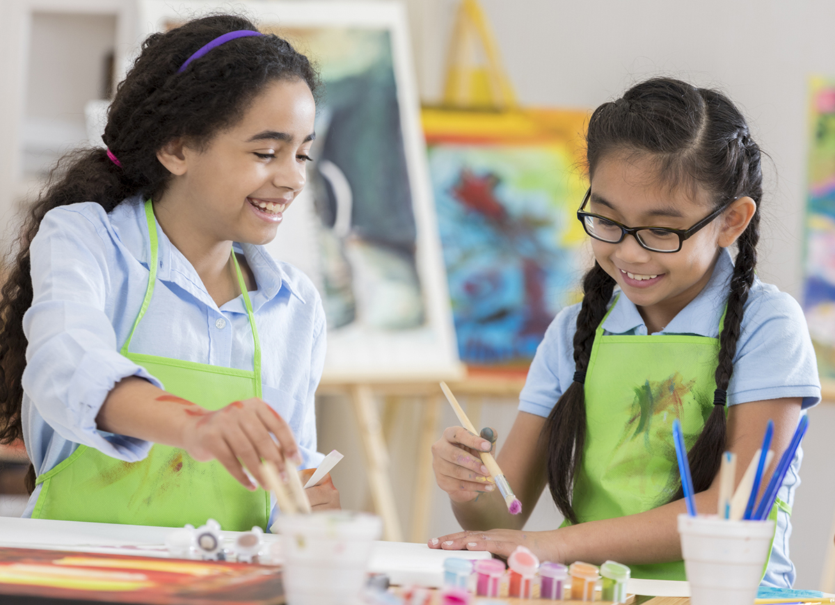 Asian and African American girls paint together during an art lesson. They are wearing aprons.
