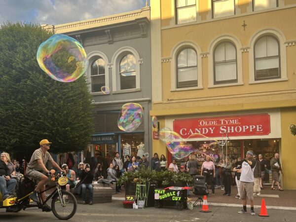 Exterior of Olde Tyme Candy Shop, with bright red sign against yellow building. In front, kids play with large bubbles.