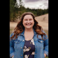 Headshot of a woman with brown wavy hair, wearing a navy floral blouse and jean jacket.